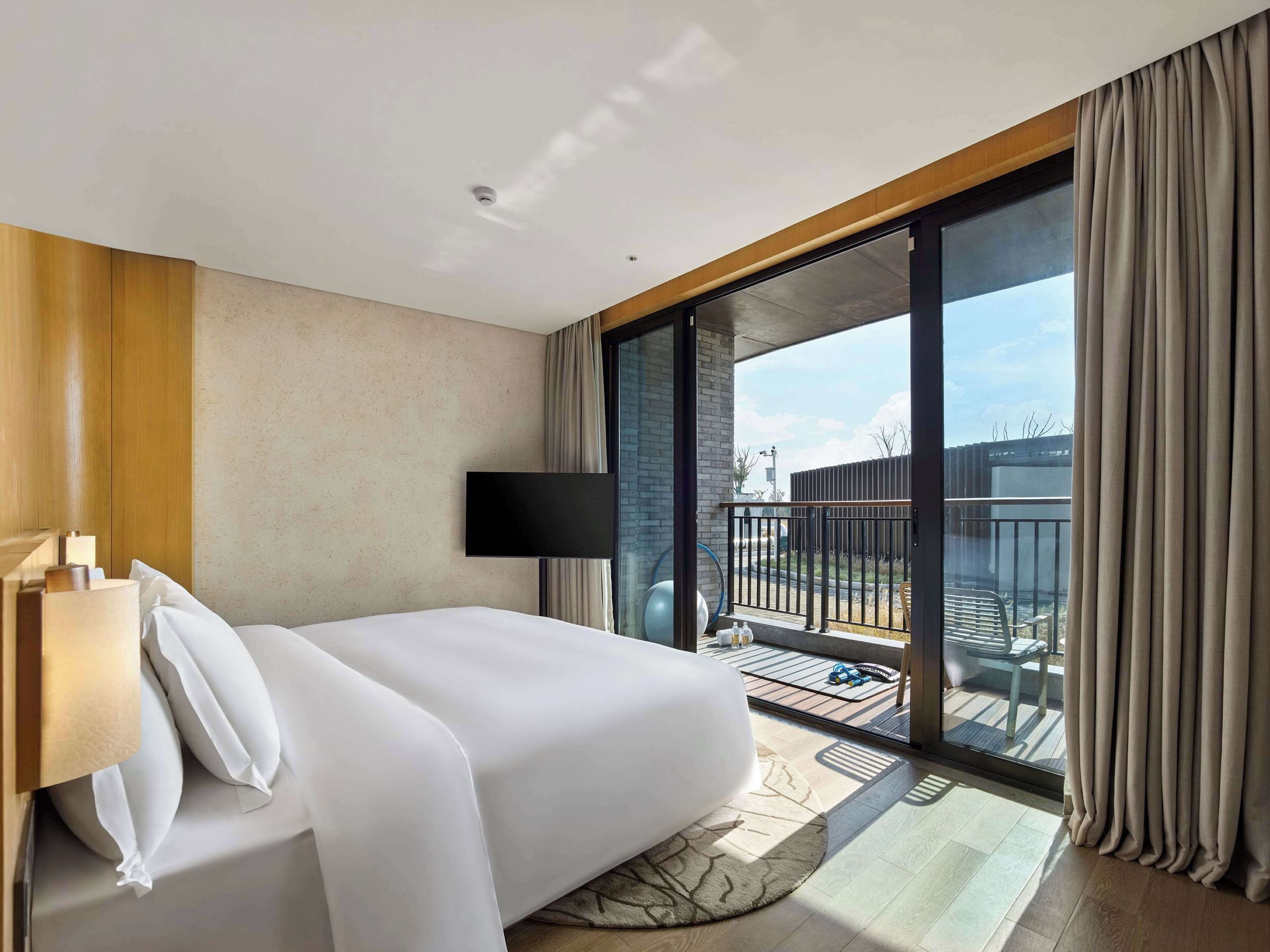 A hotel room with a bed, a flat-screen TV, a balcony with a view, and a wooden floor.