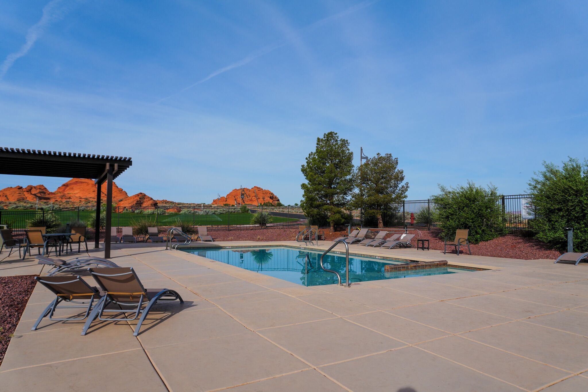 A swimming pool with lounge chairs and a covered seating area, surrounded by a fence and desert landscape.