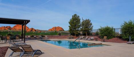 A swimming pool with lounge chairs and a covered seating area, surrounded by a fence and desert landscape.