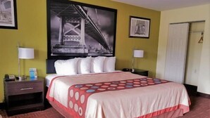 A hotel room with a bed, two bedside tables, a nightstand, a lamp, a framed picture on the wall, and a door. - Royal Inn (Bellmawr)