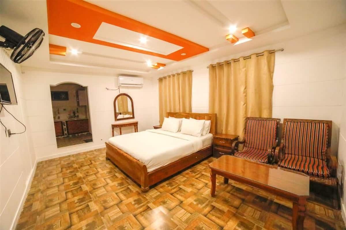 Une chambre avec un plancher en bois, un lit recouvert de draps blancs, une table de chevet en bois, un miroir, une chaise en bois et un ventilateur de plafond.