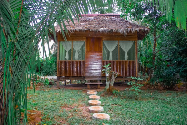 A wooden cabin with a thatched roof, surrounded by lush greenery and a stone pathway leading to the entrance.