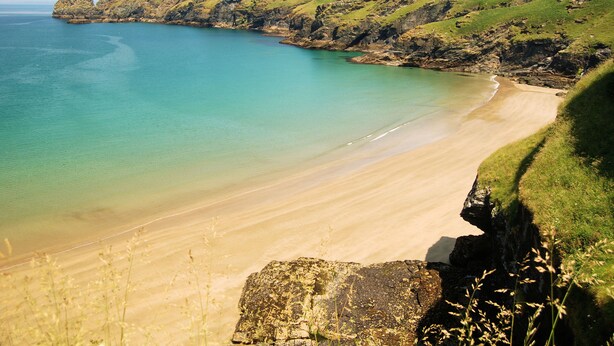A coastal landscape with a sandy beach, grassy cliffs, and a rocky outcrop. - Crowsnest - Overlooking Trebarwith Strand (Tintagel)