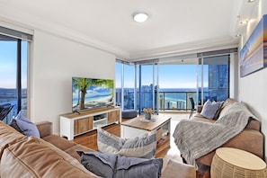 Relax in a spacious living room with a sofa set, coffee table, and flat-screen TV, offering a stunning view of the city skyline and ocean.