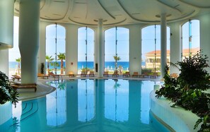 Piscina interior con grandes ventanales que dan al océano, rodeada de sillas de descanso y palmeras.