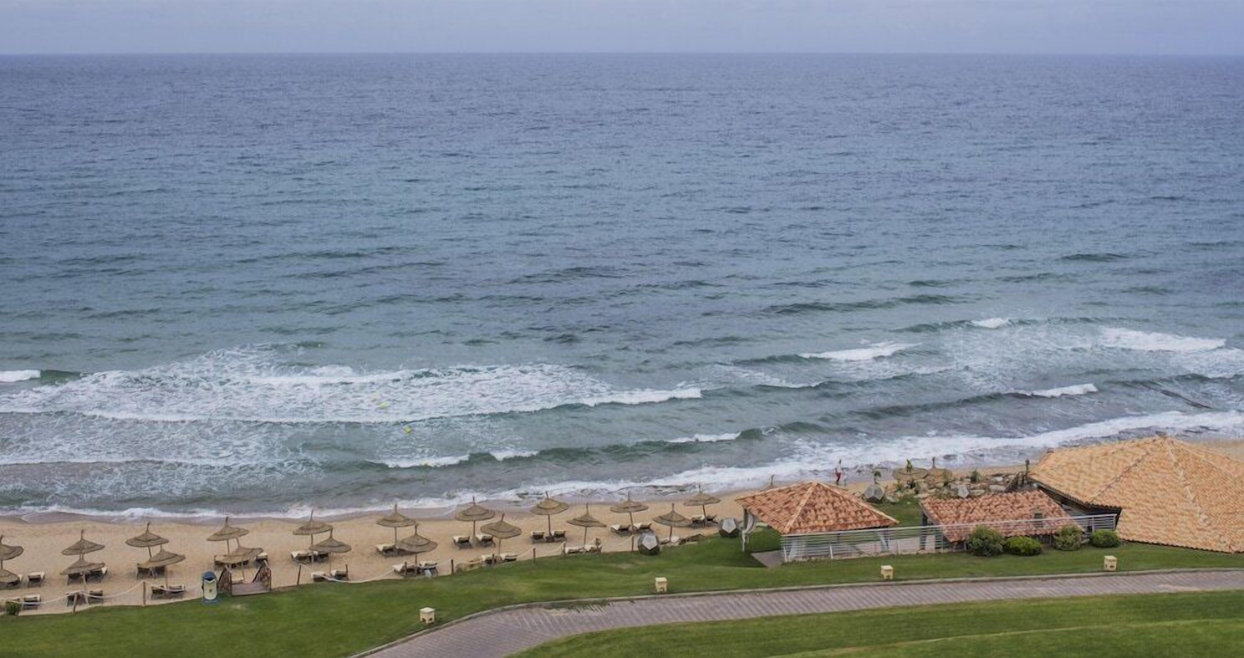 Una playa con sombrillas y sillas de descanso, un área con césped y un edificio con tejado de baldosas.