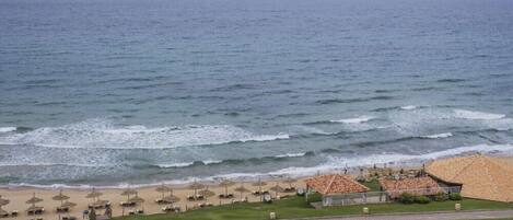 Una playa con sombrillas y sillas de descanso, un área con césped y un edificio con tejado de baldosas.