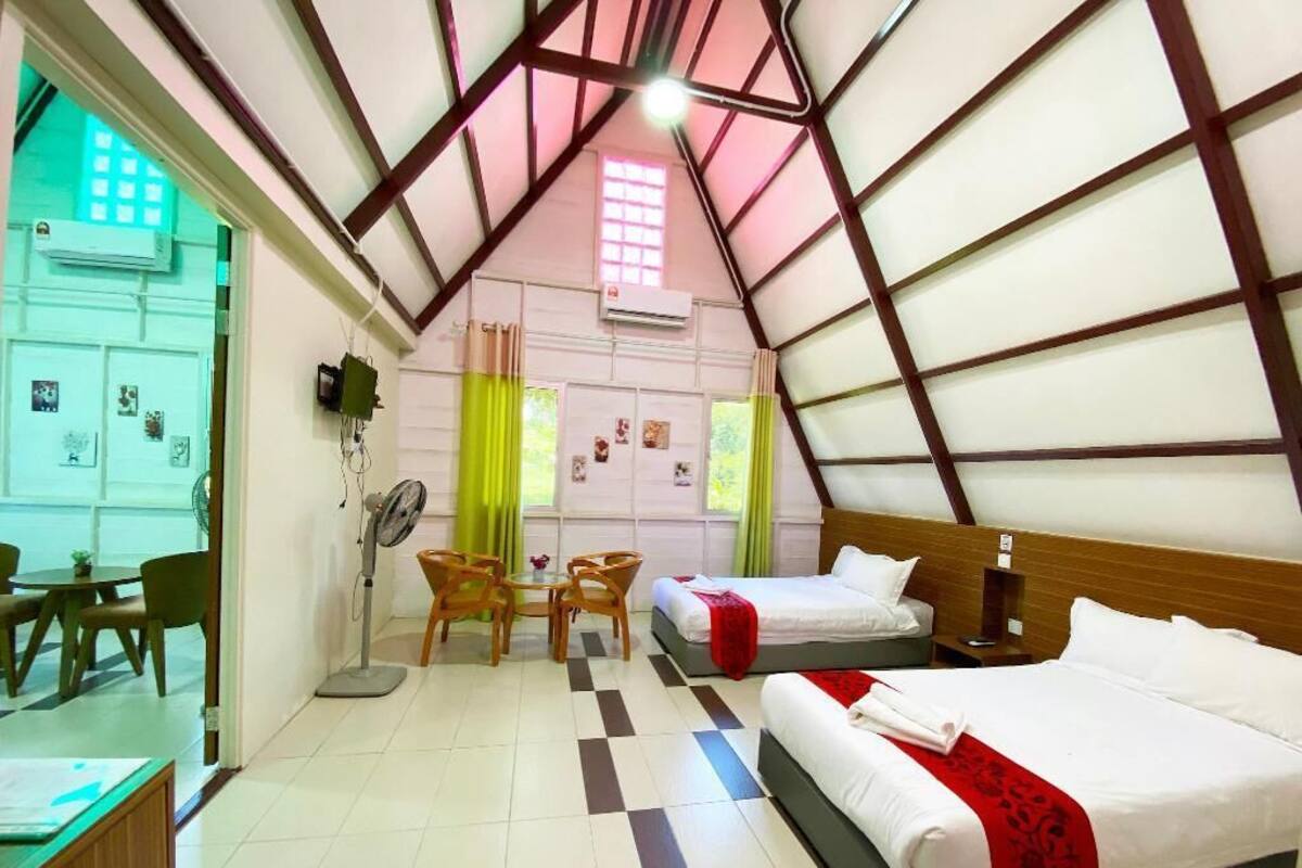 Ein Zimmer mit zwei Betten, einem Esstisch, Stühlen, einem Ventilator und einem Fenster mit Vorhängen.