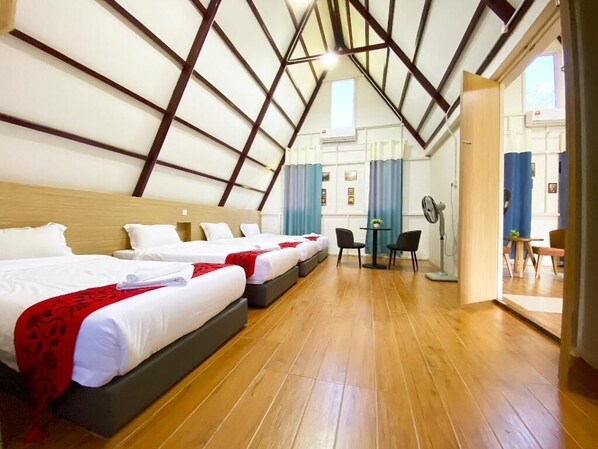 A hotel room with three beds, a wooden floor, a dining area with a table and chairs, and a view of the outside through a door.