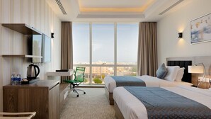 A hotel room with two beds, a desk with a kettle, a chair, and a large window offering a city view. - The Proud Hotel (Al Khobar)