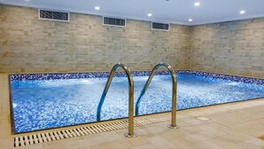 An indoor swimming pool with clear blue water, a tiled floor, and a stone wall. - The Proud Hotel (Al Khobar)