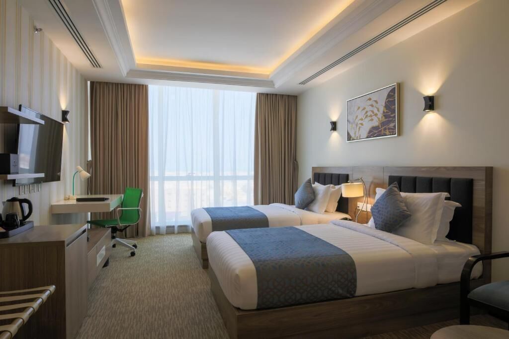 A hotel room with two beds, a desk, a chair, a TV, and a window with curtains.