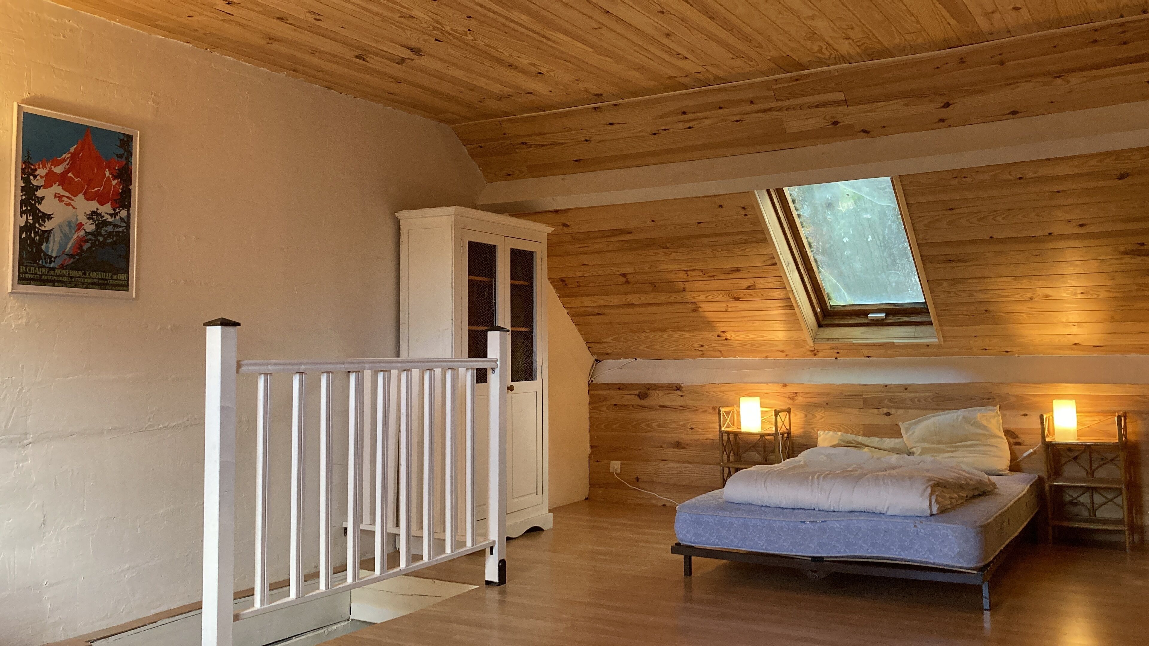A cozy attic bedroom with a sloped ceiling, a bed, a small table with a candle, and a window providing natural light.