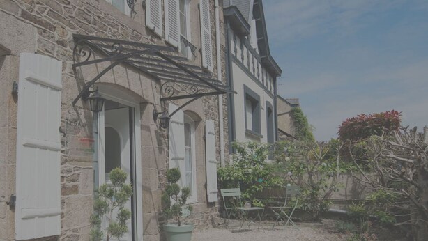 Un edificio de piedra con un estilo arquitectónico europeo tradicional, que presenta fachada de piedra, contraventanas blancas y un pequeño jardín con plantas en macetas y un banco.