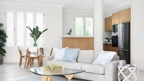Ein modernes Wohnzimmer mit Glastisch, einer Couch mit Kissen, einem Essbereich mit Stühlen und einer Küche mit Holzschränken und einem Kühlschrank.