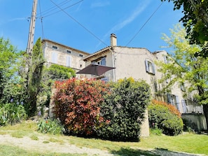 Un bâtiment en pierre doté d’une cheminée, entouré d’une végétation luxuriante et surplombé d’un ciel bleu clair.