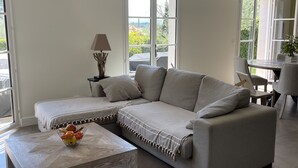 Un moderno salón con un sofá modular, una mesa de centro con fruta, una lámpara y grandes ventanales que ofrecen vistas a zonas verdes.