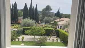Vista a través de una ventana que muestra un jardín con un columpio, setos bien cuidados y árboles altos.