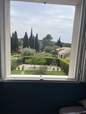 Une vue à travers une fenêtre sur un jardin avec une balançoire, des haies soigneusement taillées et de grands arbres.