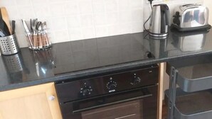 Cozinha com bancada preta, torradeira de aço inoxidável e cafeteira. Há utensílios em um suporte e um forno preto abaixo da bancada.
