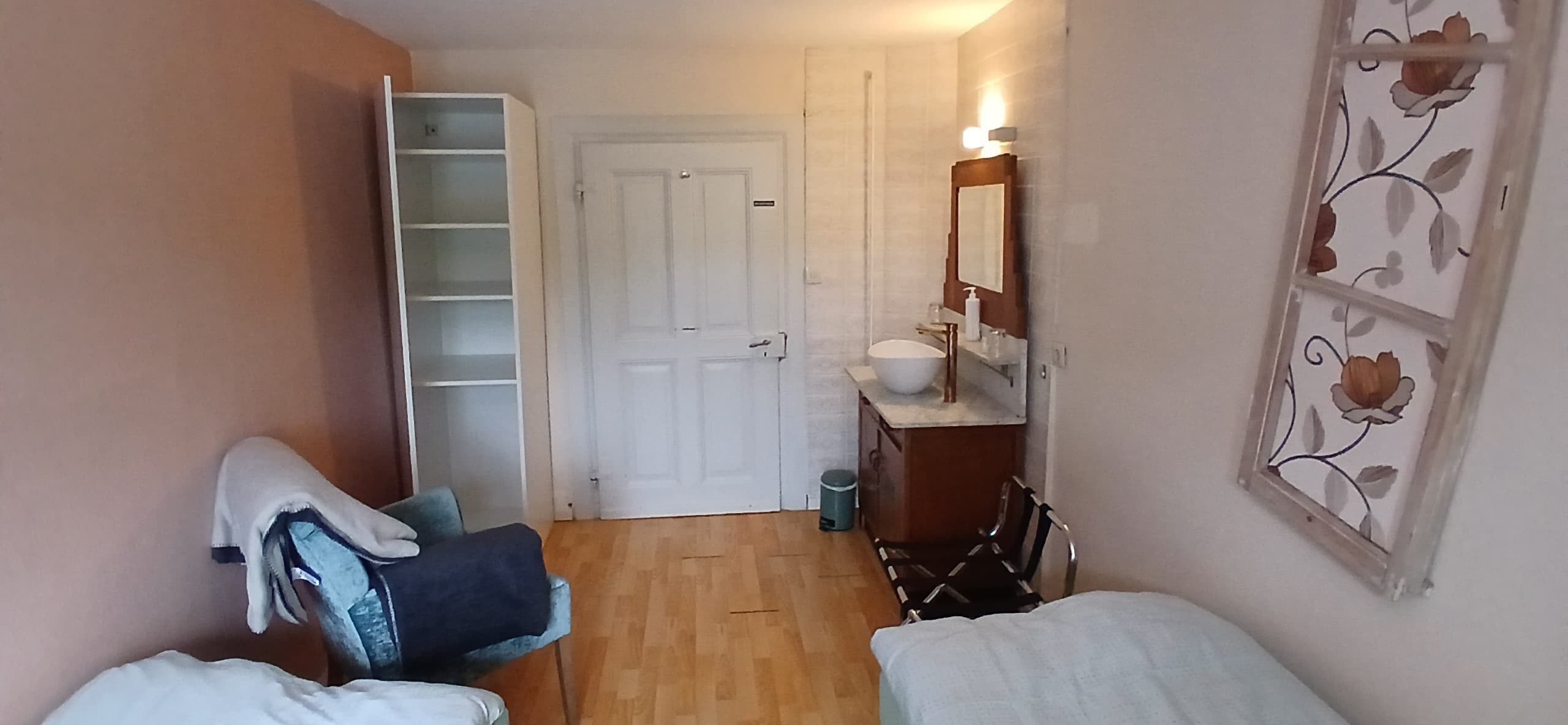 A small room with a bed, a chair, a wardrobe, and a bathroom with a sink and mirror.
