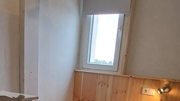 A small, cozy room with a bed, a chair, a window, and a wall-mounted shelf.