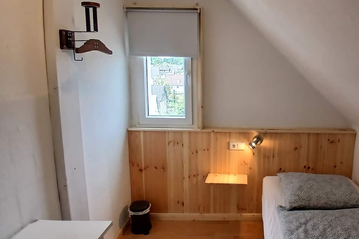 A small, cozy room with a sloped ceiling, a single bed with a gray blanket, a small window with a view of greenery, a wall-mounted light, and a wooden panel wall.
