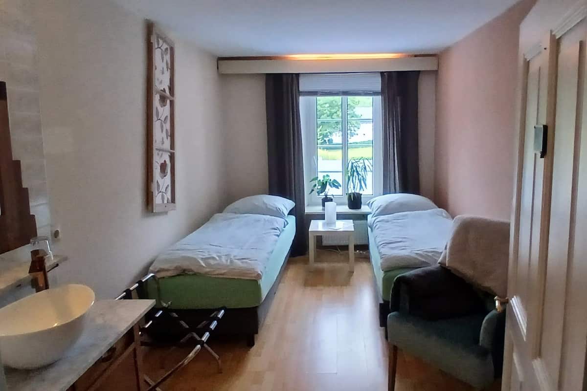 A narrow room with two single beds, a small table, a sink, and a window with a view of greenery.
