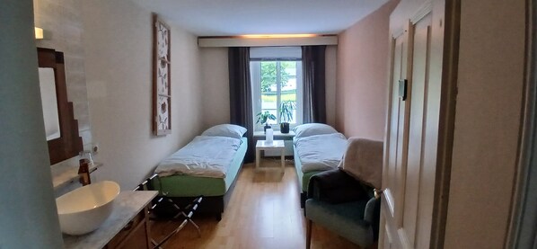 Habitación pequeña con dos camas individuales, una mesita, un lavamanos y una ventana que da a zonas verdes.