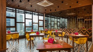 Ein modernes Restaurant mit Holzbalkendecken, großen Fenstern mit Blick auf die Stadt, gelben Stühlen und Tischen mit roten Untersetzern.
