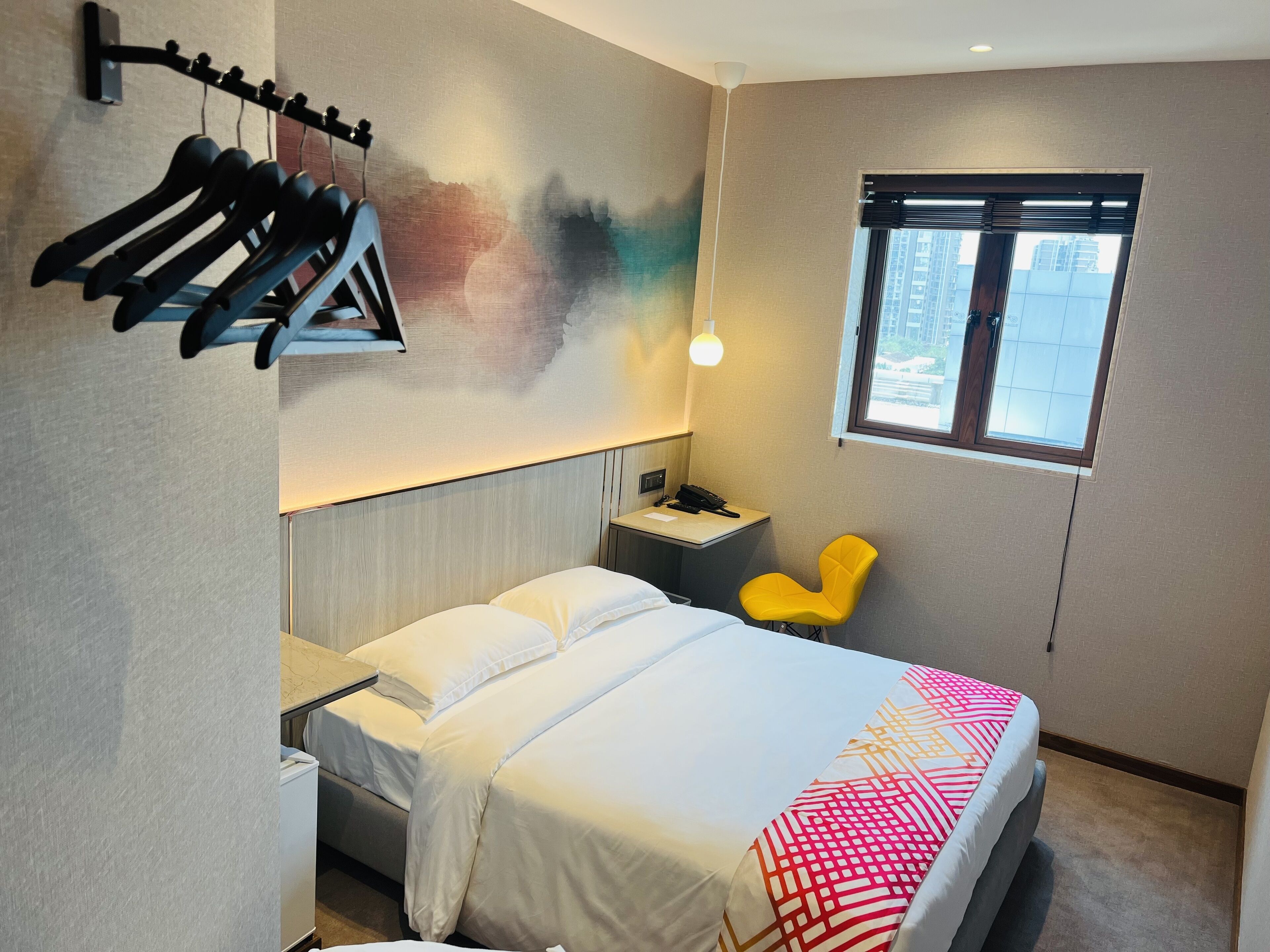 A hotel room with a bed, a chair, a desk with a phone, a window, and a wall-mounted rack with hangers.