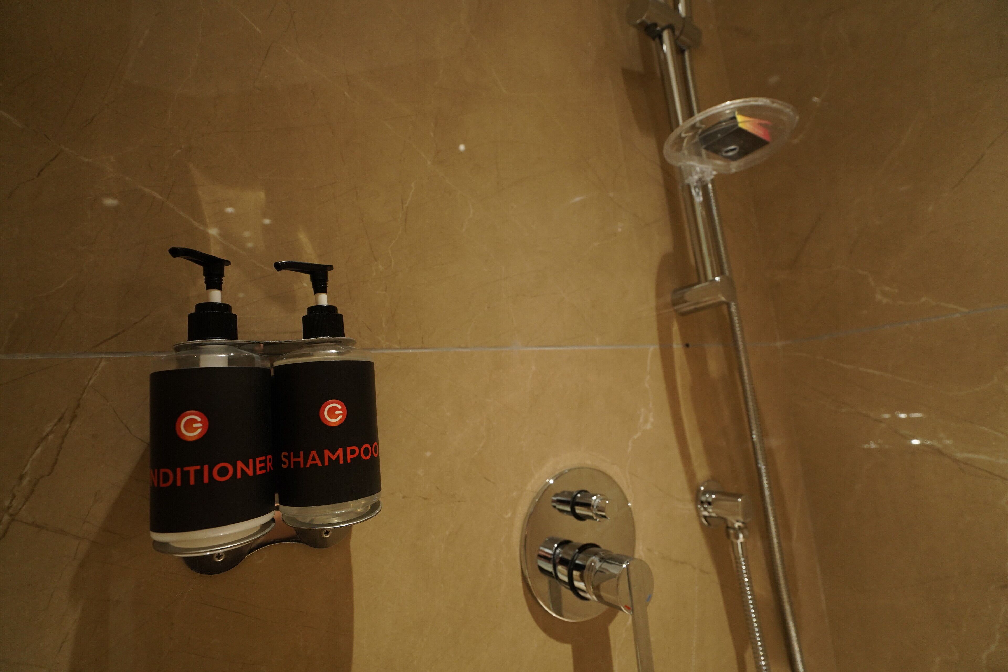 Un baño con piso de mármol, dos botellas de acondicionador y champú, y una ducha con cabezal de ducha manual.