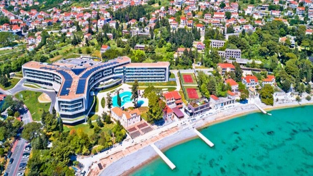 An aerial view of a coastal town with a modern hotel featuring a unique curved architecture, a beach, and a marina.