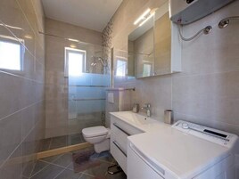 A modern bathroom with a glass shower enclosure, a white toilet, a double sink vanity, and a built-in washing machine.