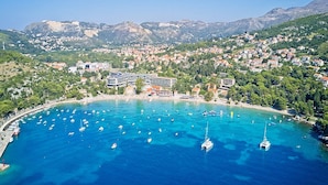 An aerial view of a coastal town with a marina, a prominent hotel with a curved facade, and surrounding hills.