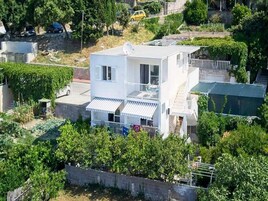 Un edificio blanco de dos pisos con un balcón, rodeado de vegetación y un muro de piedra.