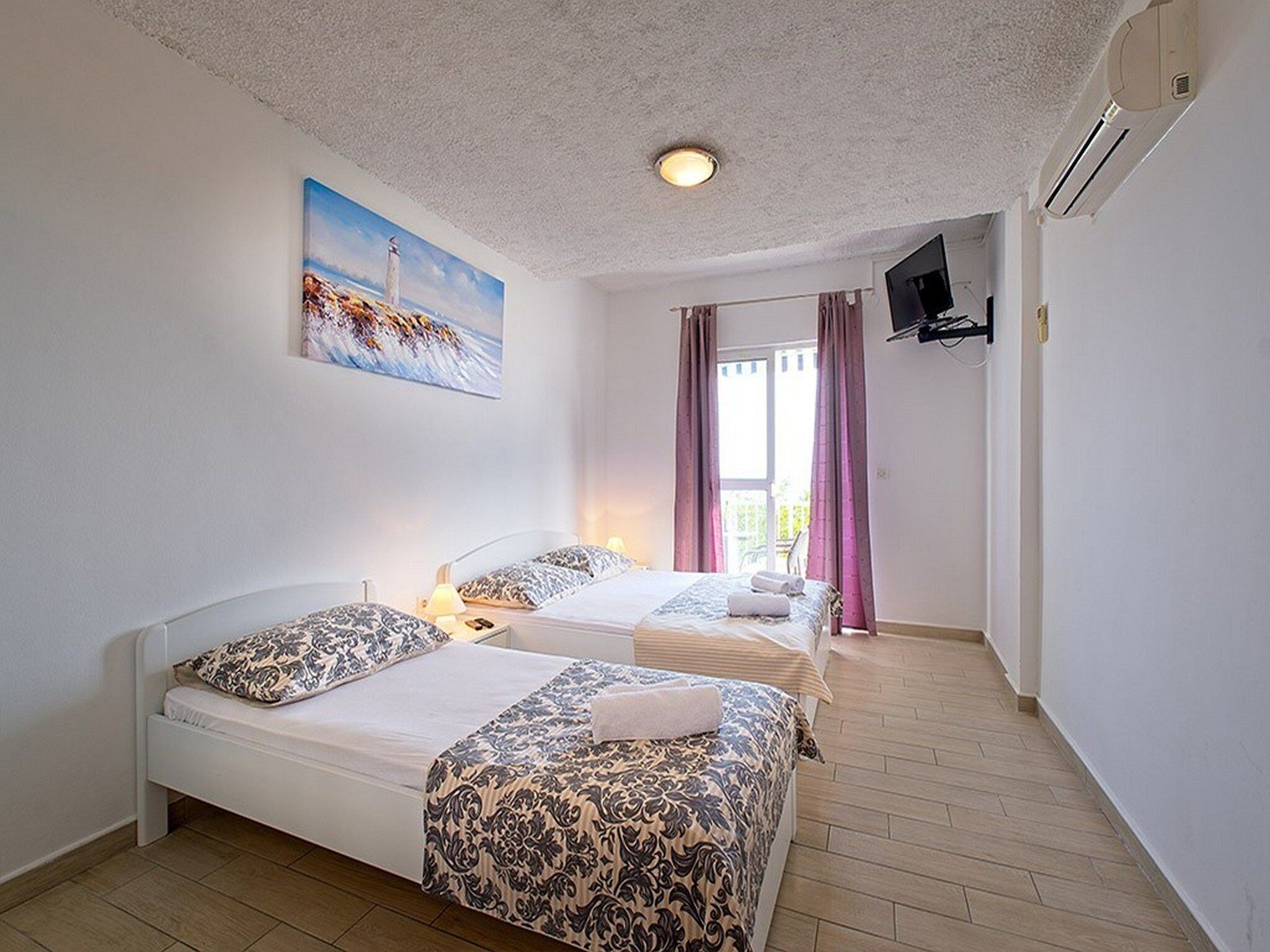 A hotel room with two beds, a painting of a lighthouse, a TV mounted on the wall, and a window with curtains.