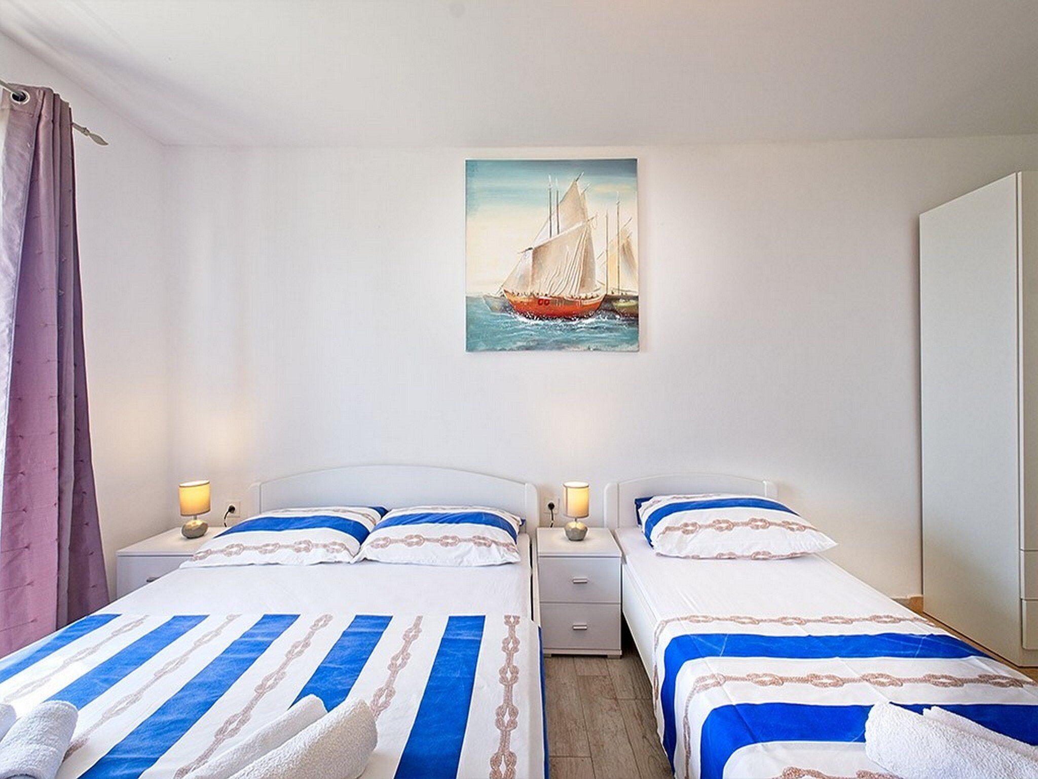 Ein Hotelzimmer mit zwei Betten, jeweils mit blau-weißer Bettwäsche, einem Segelbootgemälde an der Wand und einem Fenster mit Vorhängen.