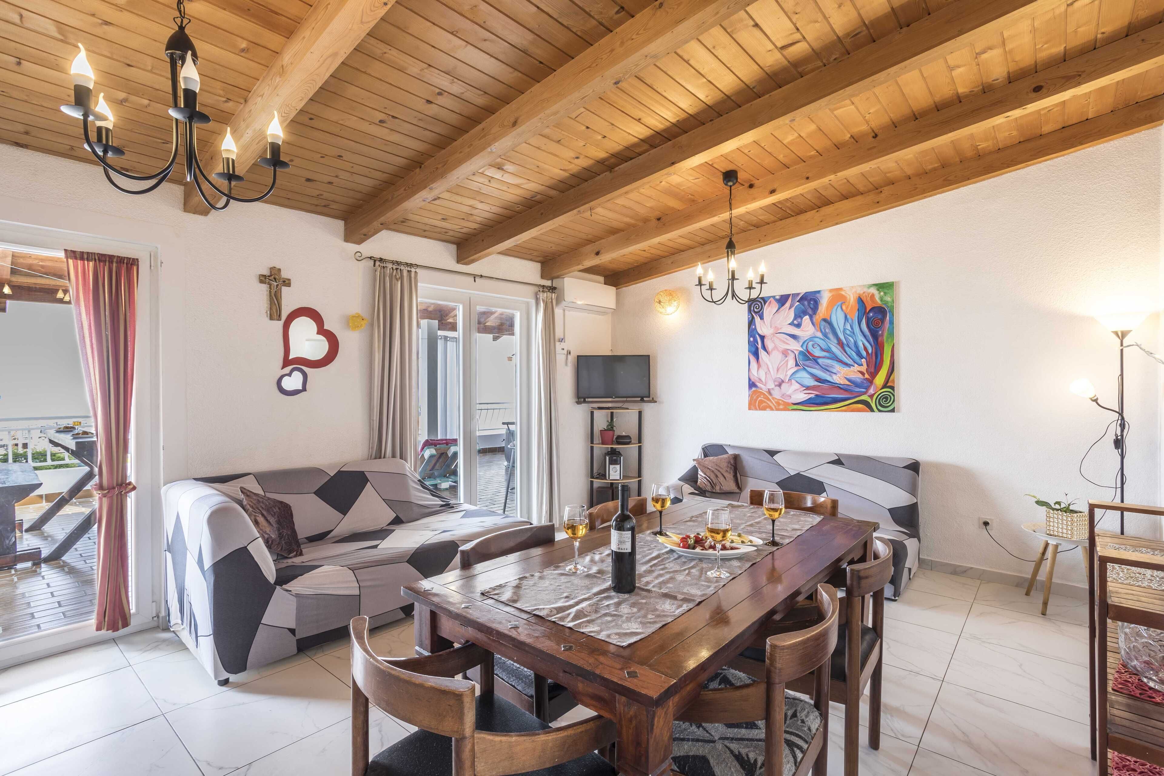 Un comedor con una mesa de madera, sillas y una lámpara de araña. Se observa un sofá y un televisor al fondo.