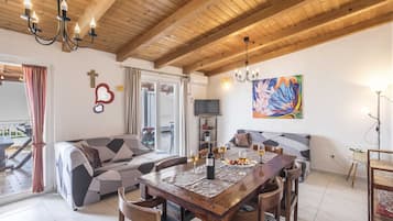 Un comedor con una mesa de madera, sillas y una lámpara de araña. Se observa un sofá y un televisor al fondo.