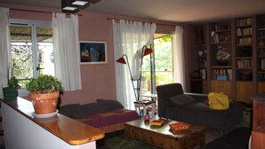 Een gezellige woonkamer met een bank, fauteuil en een salontafel. Er is een boekenplank vol boeken en een plant in een pot op het raamkozijn.
