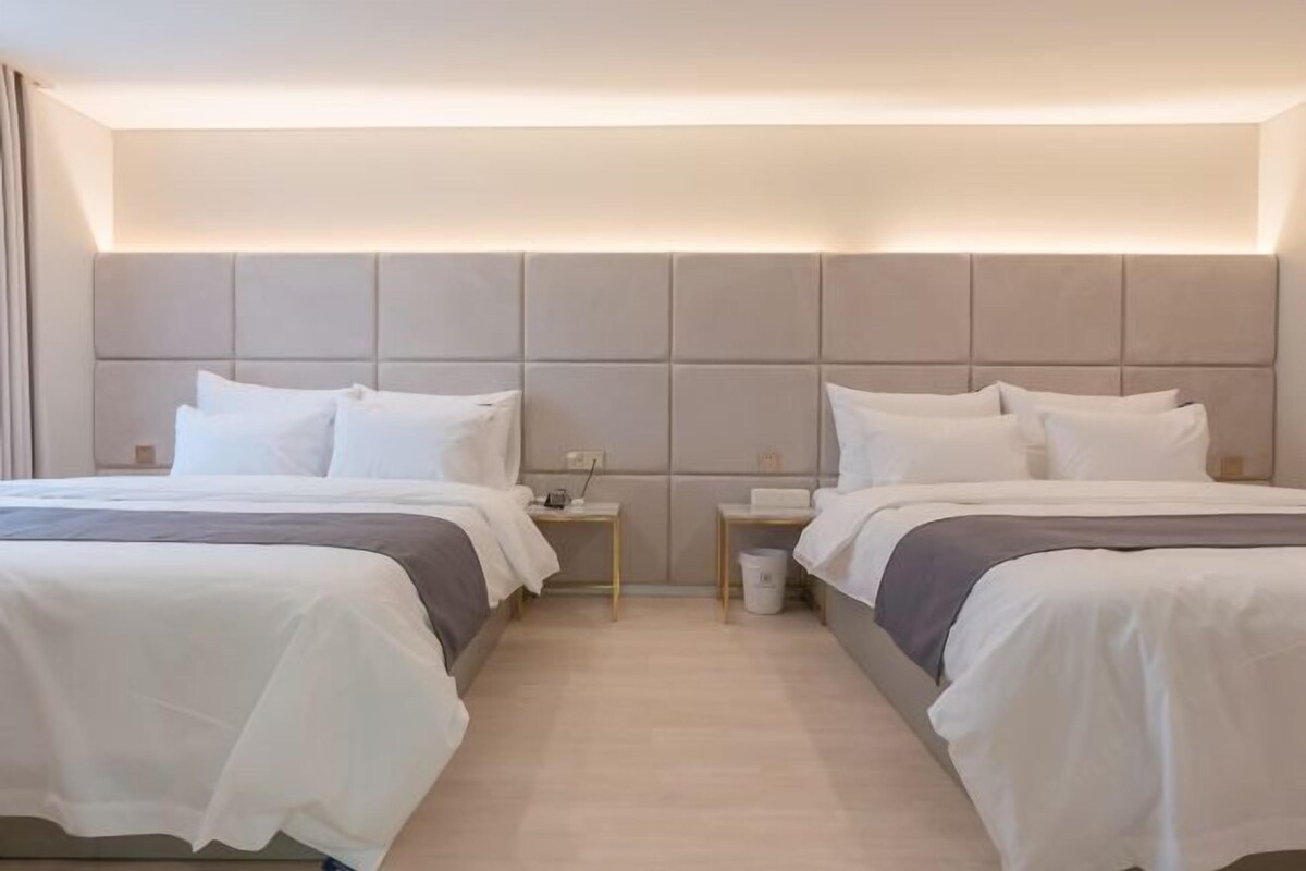 Habitación de hotel con dos camas, un cabecero grande acolchado, mesitas de noche y un cubo de basura.
