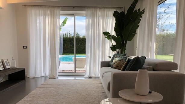 A modern living room with a sofa, a coffee table, and a large plant. There is a sliding glass door leading to a pool area. - Apartment (Pietrasanta)