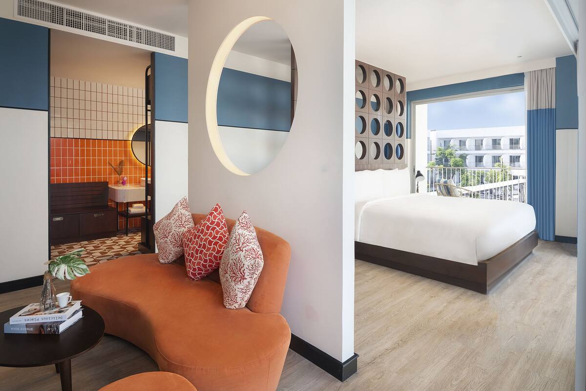 Ein modernes Hotelzimmer mit einem großen Bett, einer Sitzecke, einem runden Spiegel und Blick auf die Stadt durch große Fenster.