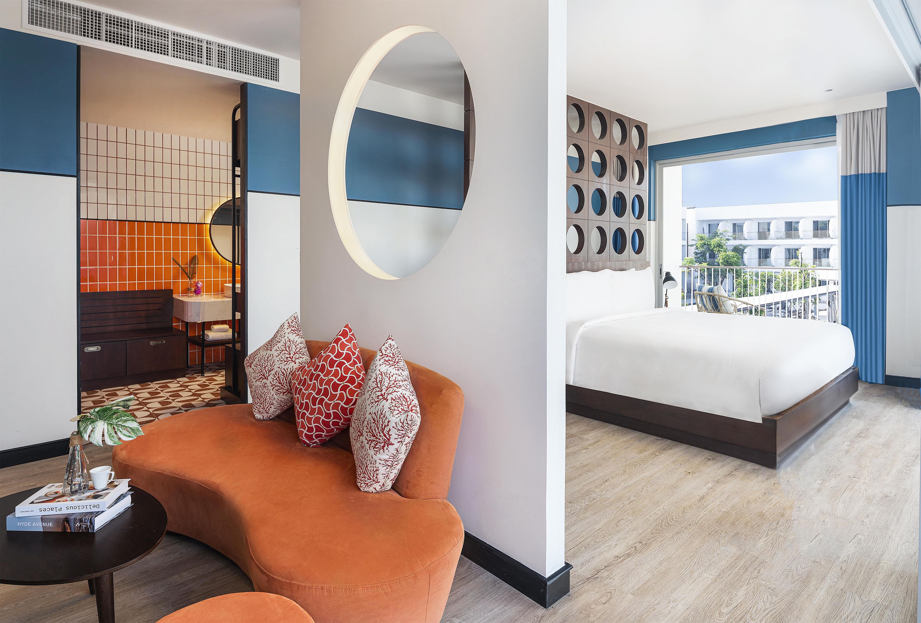 A modern hotel room with a large bed, a sofa with decorative pillows, a round mirror, and a view of the city.
