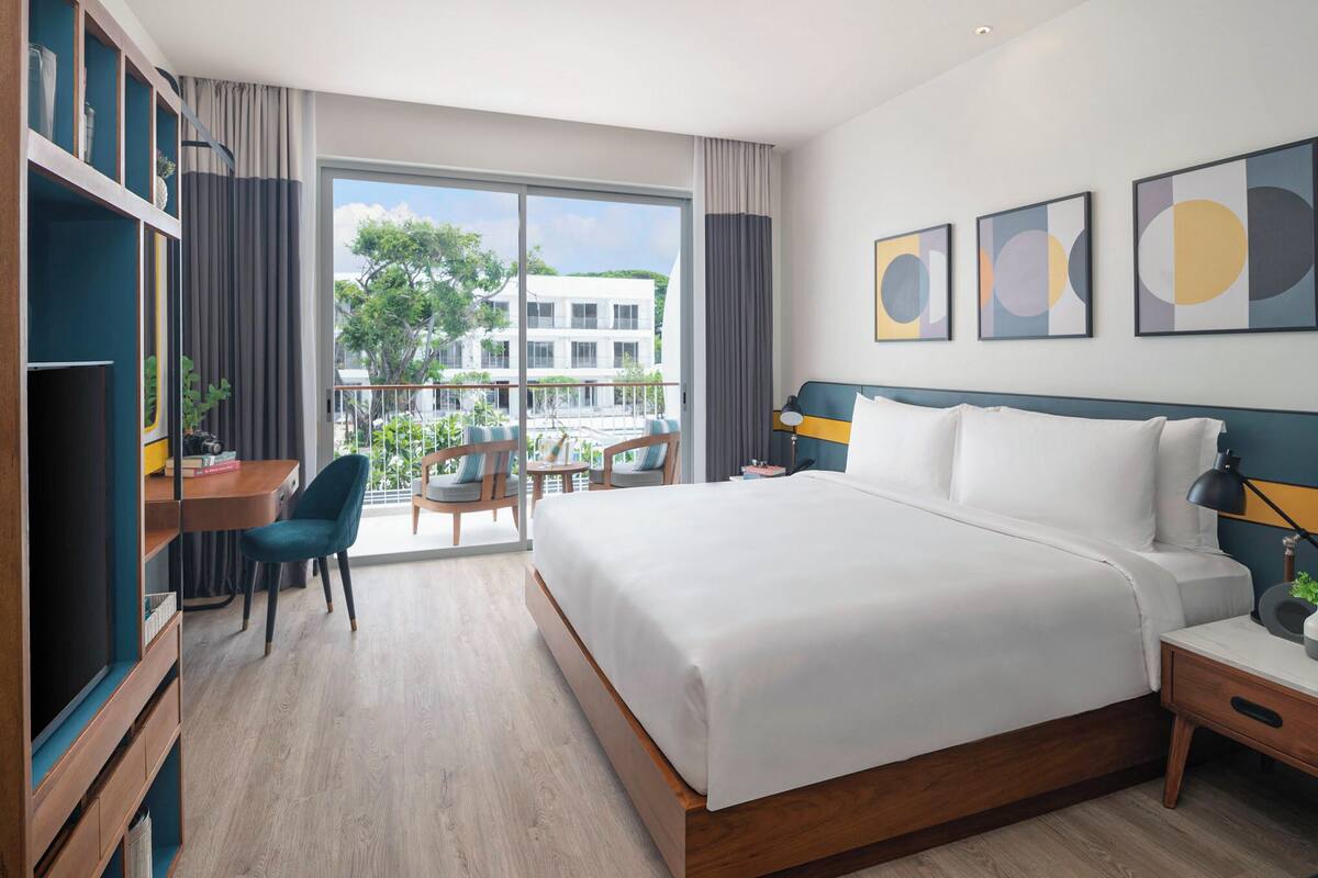 Ein modernes Hotelzimmer mit einem großen Bett, einem Schreibtisch mit Stuhl und einem Balkon mit Blick auf Gebäude und Bäume.