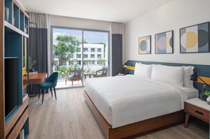 A modern hotel room with a large bed, a desk with a chair, and a balcony with a view of buildings and trees.