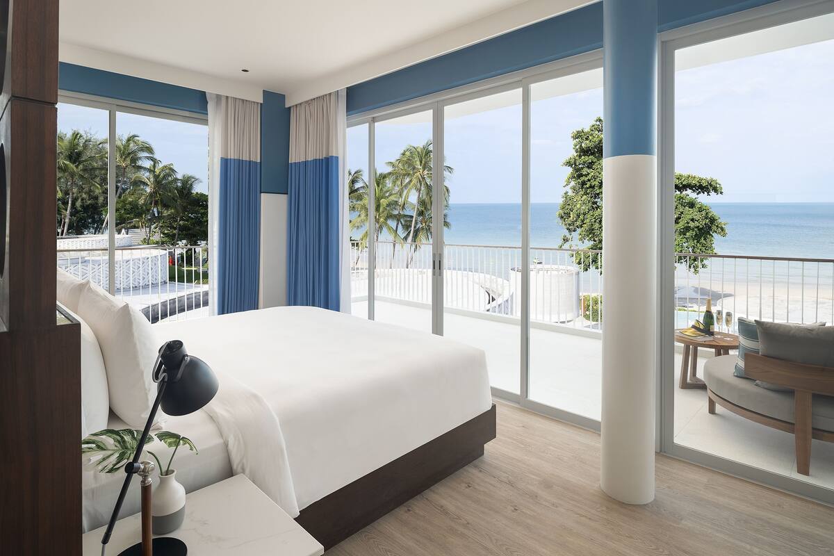 Ein modernes Hotelzimmer mit einem großen Bett, einem Balkon mit Blick auf den Strand und einer Sitzecke mit Tisch und Stühlen.