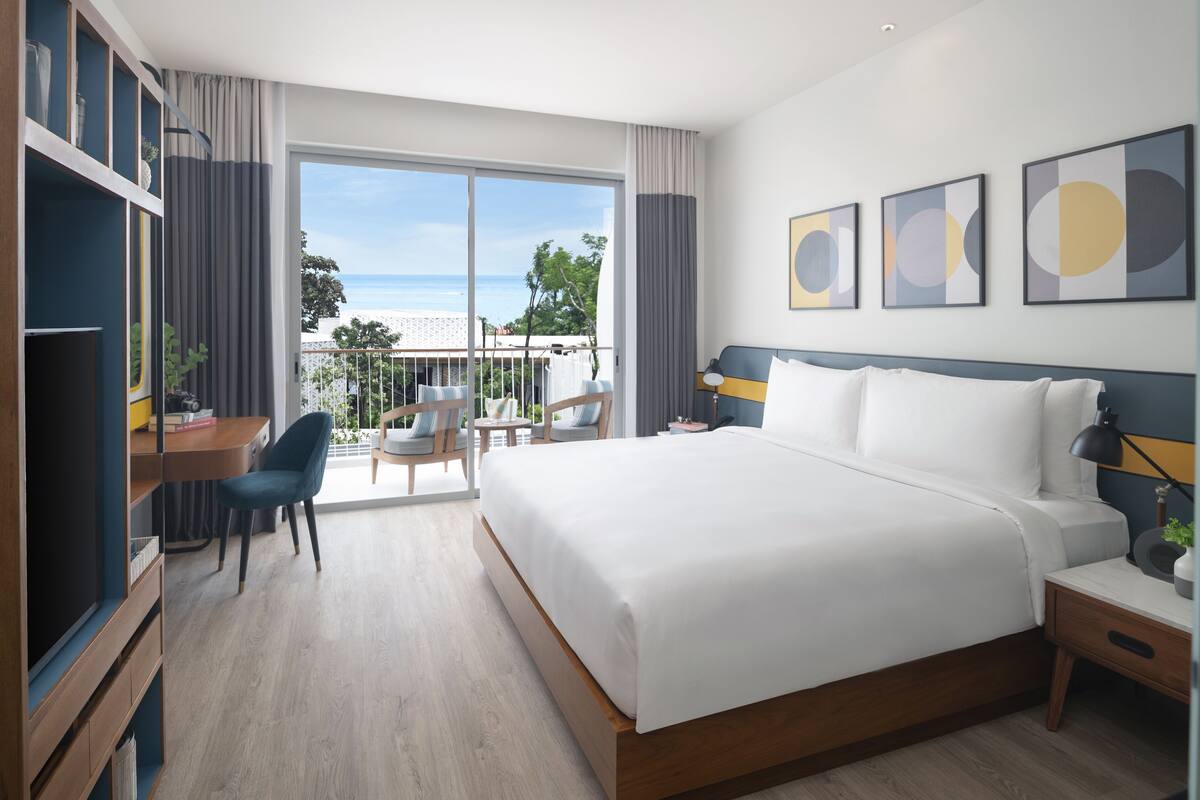Ein modernes Hotelzimmer mit einem großen Bett, einem Schreibtisch mit Stuhl und einem Balkon mit Meerblick.