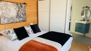 Un lit bien fait, avec un oreiller à motifs noir et blanc, une couverture noire et un plaid orange. On peut voir une tête de lit en bois et un tableau encadré au mur.
